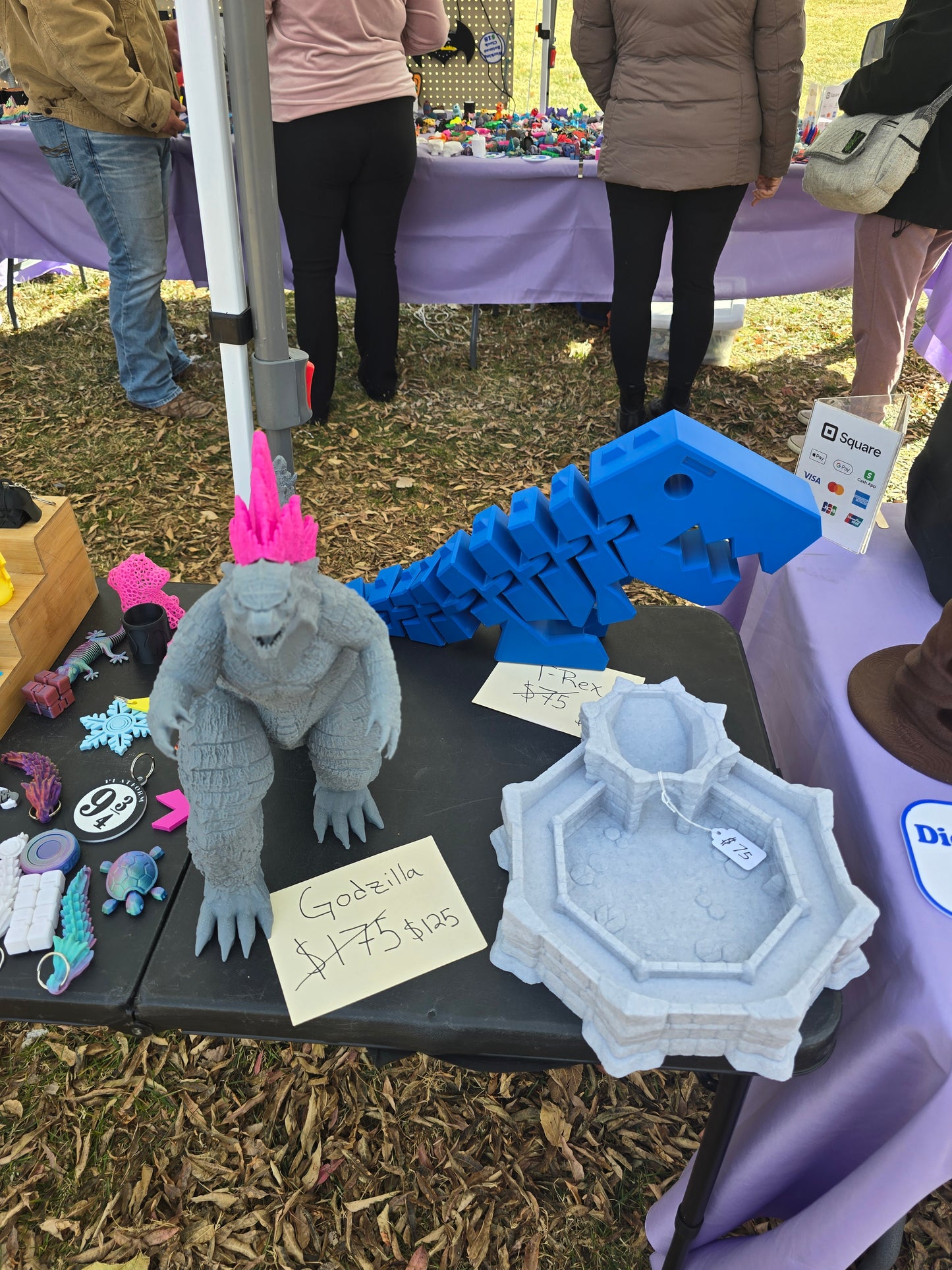 Large 3D printed grey Godzilla with pink dorsal fins on a display table at an outdoor market.