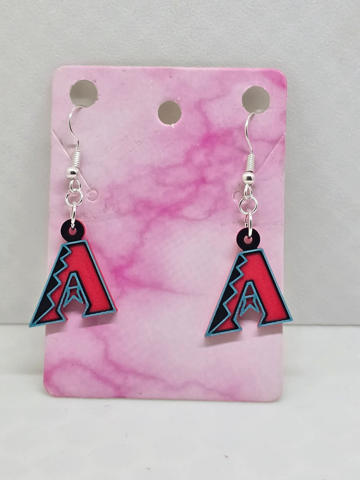 Small 3D printed Arizona Diamondbacks logo dangle earrings with black, red, and teal accents on a pink marble display card.