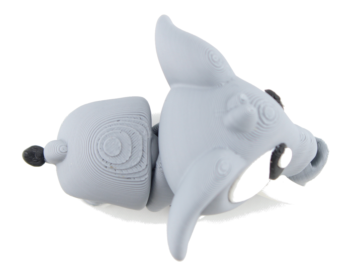Overhead view of the 3D printed gray baby elephant on its belly, ear lifted, white eye and tusk visible beside the curled trunk