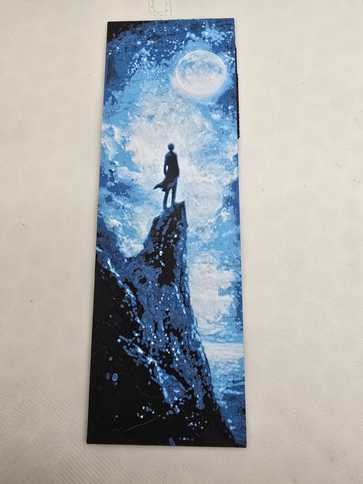 A book mark of a painting of a man standing on a cliff, gazing at a large moon illuminating the night sky.