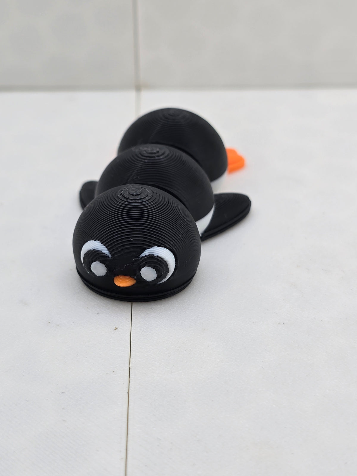 3D printed black penguin figurine with large white eyes and orange beak in a lying down pose.