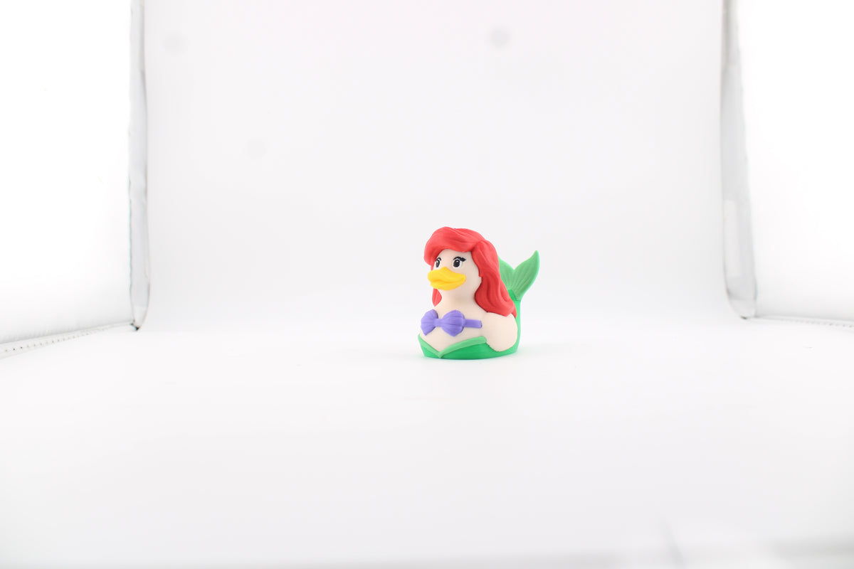 3D printed Ariel duck in a white lightbox, front three-quarter view with flowing red hair, yellow bill, purple shell top and her green tail fin raised behind her