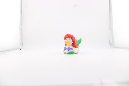 3D printed Ariel duck in a white lightbox, front three-quarter view with flowing red hair, yellow bill, purple shell top and her green tail fin raised behind her