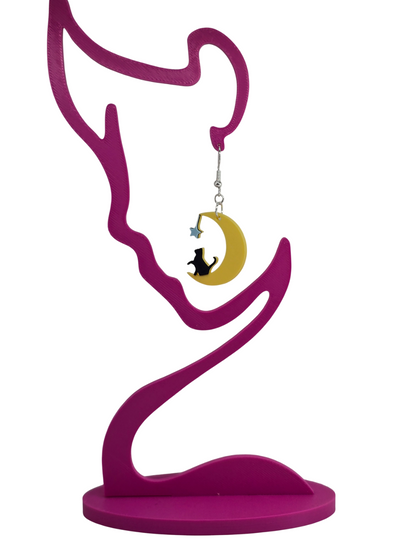 3D printed earrings featuring a black cat sitting on a yellow crescent moon with a blue star on a pink display.