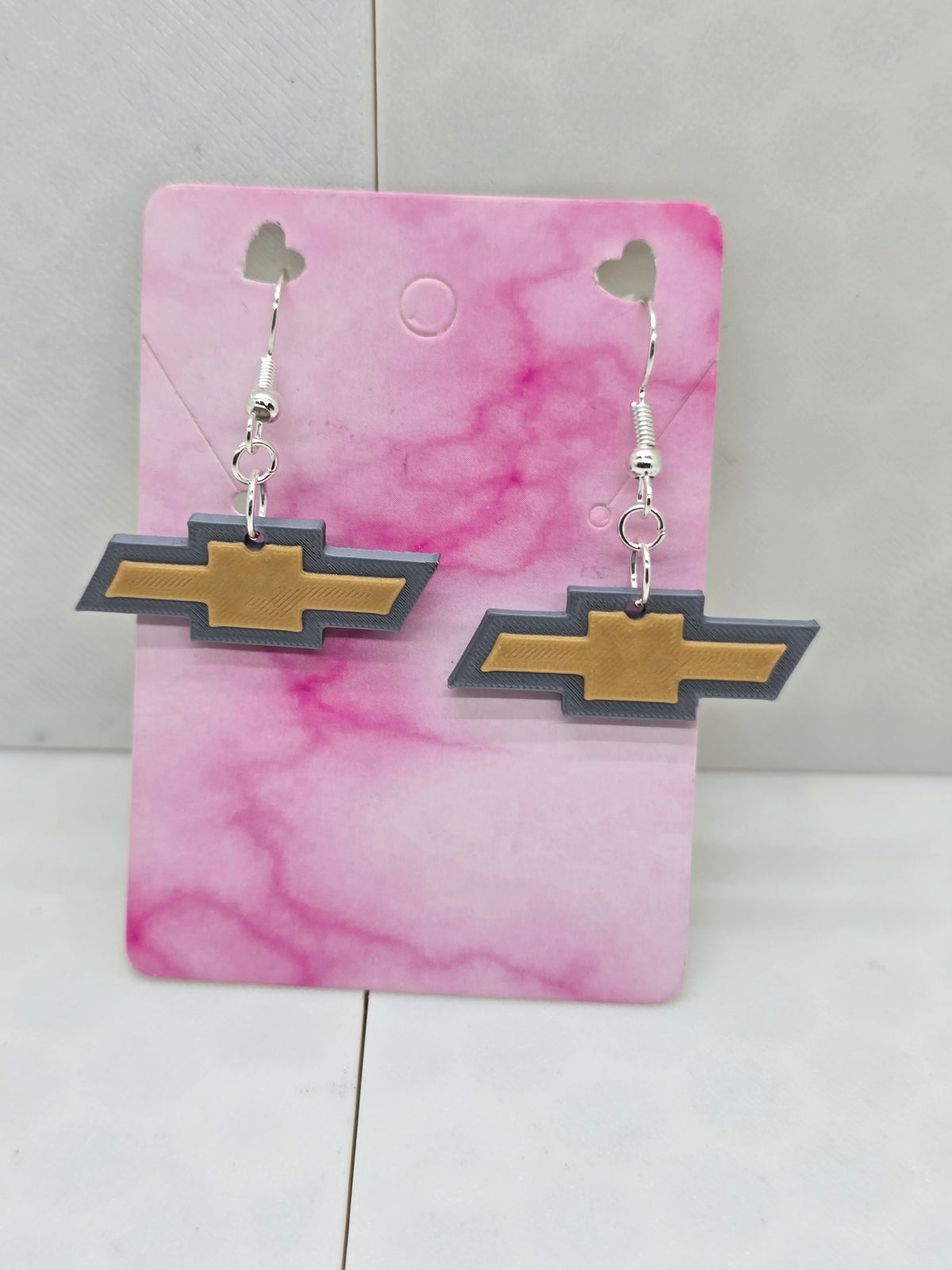 3D printed Chevy logo earrings in gold and grey on a pink marble display card.