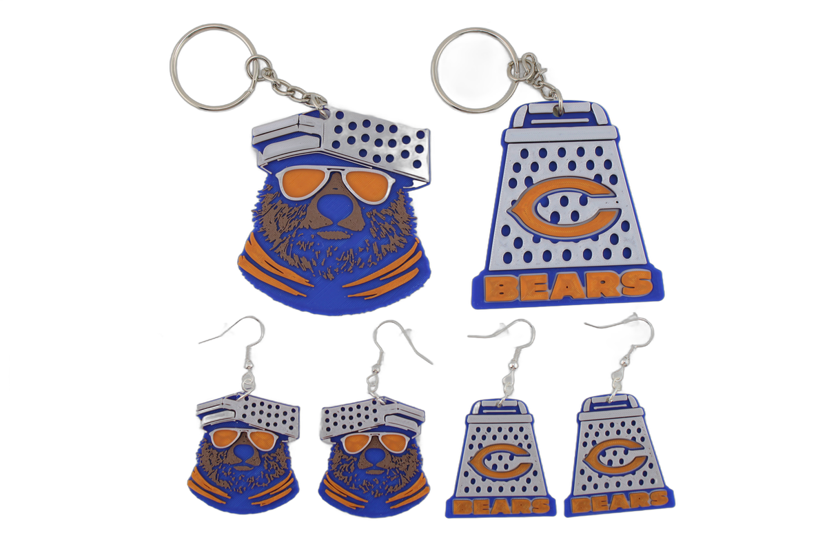 Full 3D printed Chicago Bears collection lineup — blue bear head keychain with orange sunglasses and cheese grater keychain with orange C logo on top, four cheese grater earrings below, all in Bears blue, orange, and white.