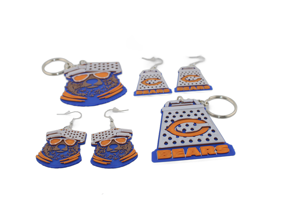 3D printed Chicago Bears earrings and keychains arranged together — blue bear heads with orange sunglasses alongside cheese graters with the orange C logo.