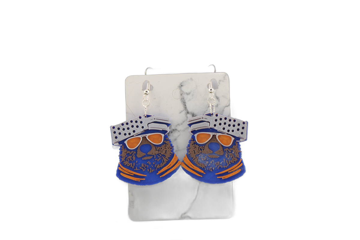 Close-up of 3D printed bear head earrings on display card — Chicago Bears blue with crisp orange sunglasses and cheese grater hat detail.