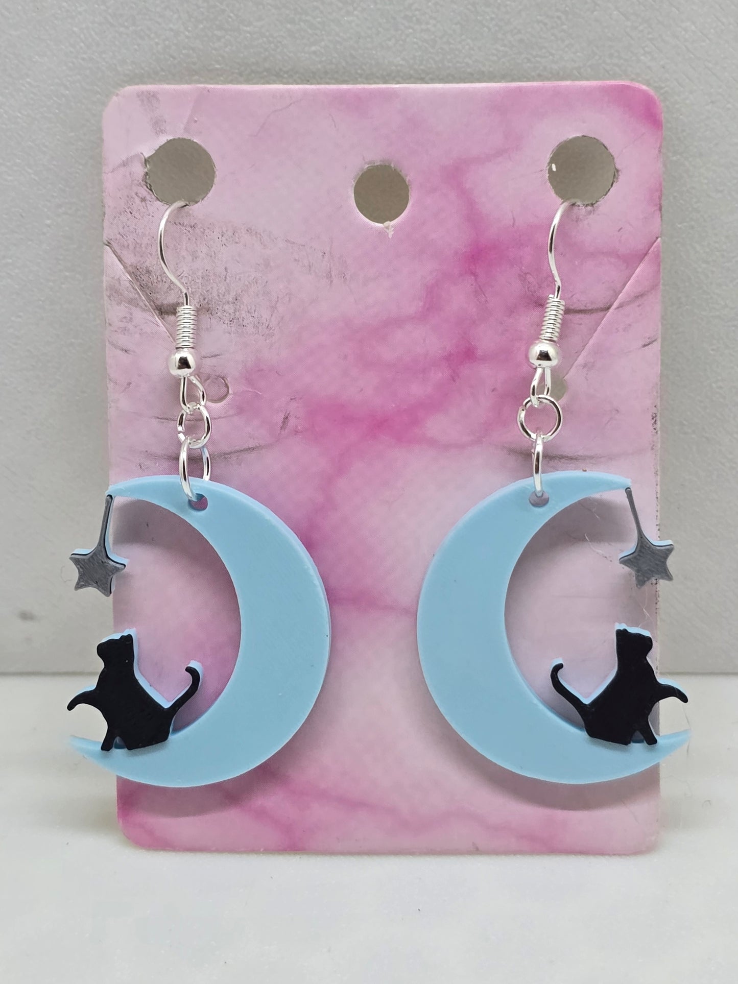 3D printed earrings featuring a black cat sitting on a light blue crescent moon with a silver star.