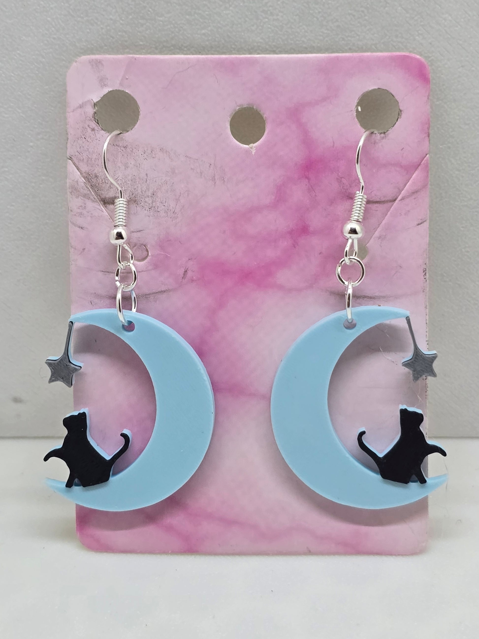 3D printed earrings featuring a black cat sitting on a light blue crescent moon with a silver star.