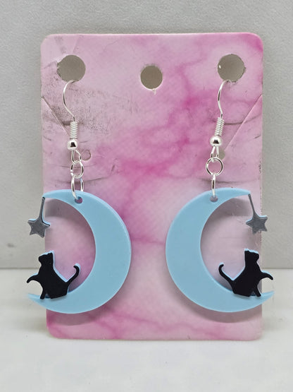 3D printed earrings featuring a black cat sitting on a light blue crescent moon with a silver star.