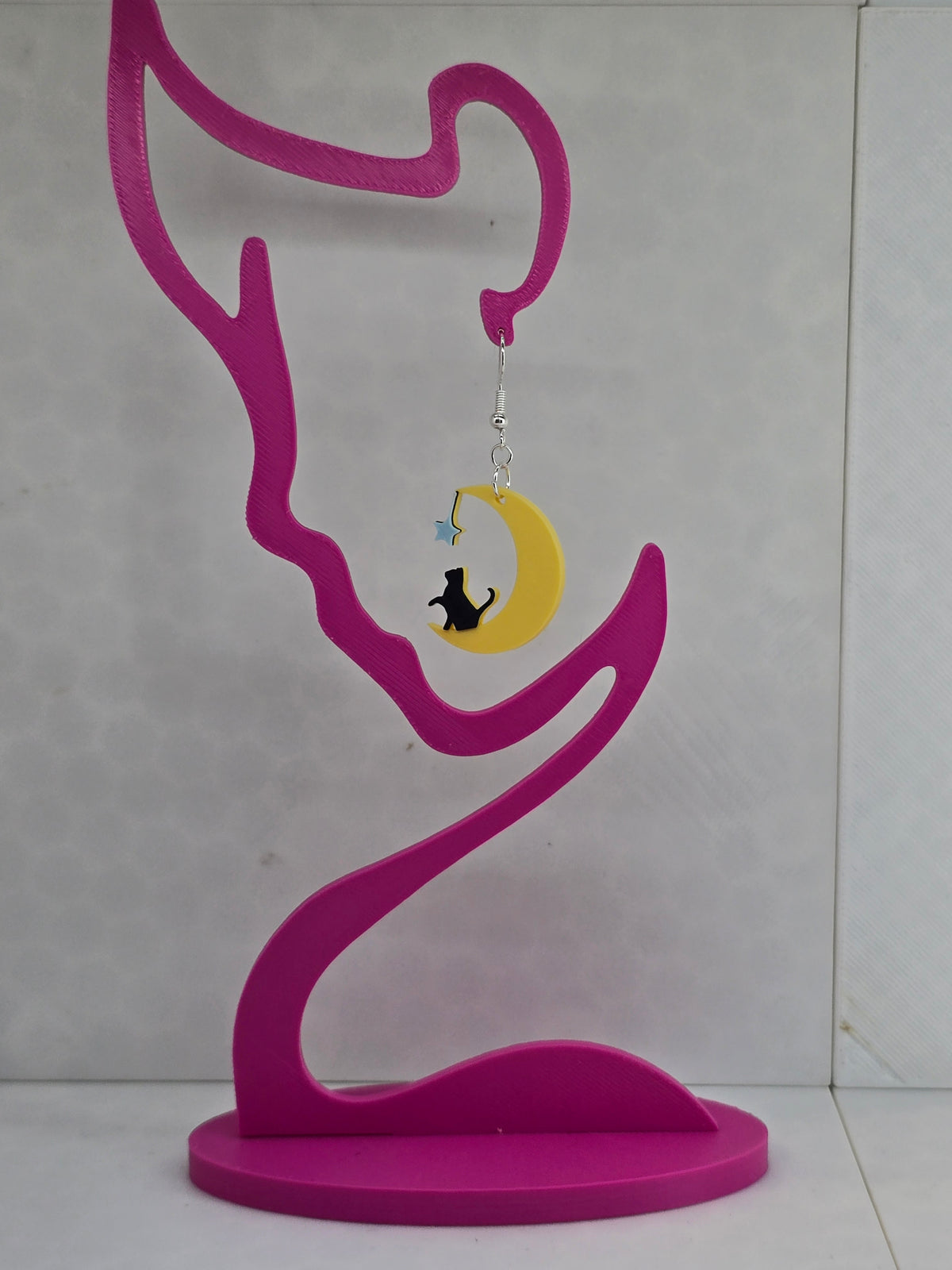 3D printed earrings featuring a black cat sitting on a yellow crescent moon with a blue star on a pink display.