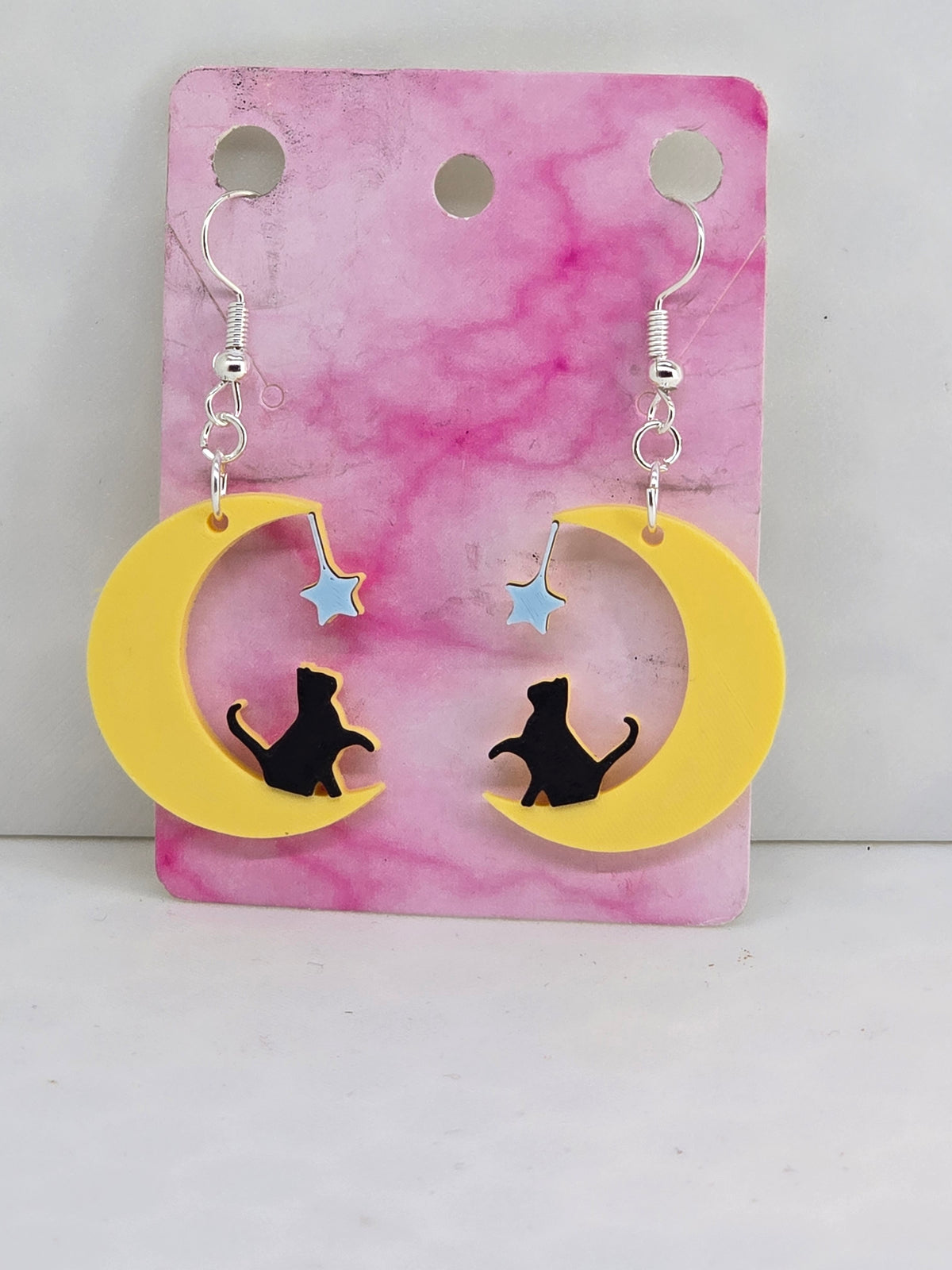 3D printed earrings featuring a black cat sitting on a yellow crescent moon with a blue star.
